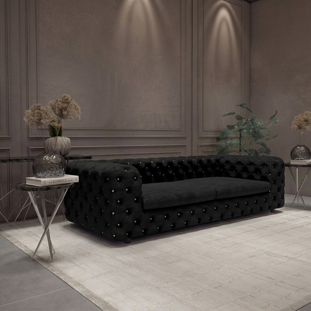 A black tufted velvet Chesterfield sofa in a modern, elegant living room setting with gray paneled walls, a light rug, and side tables with dried flowers.