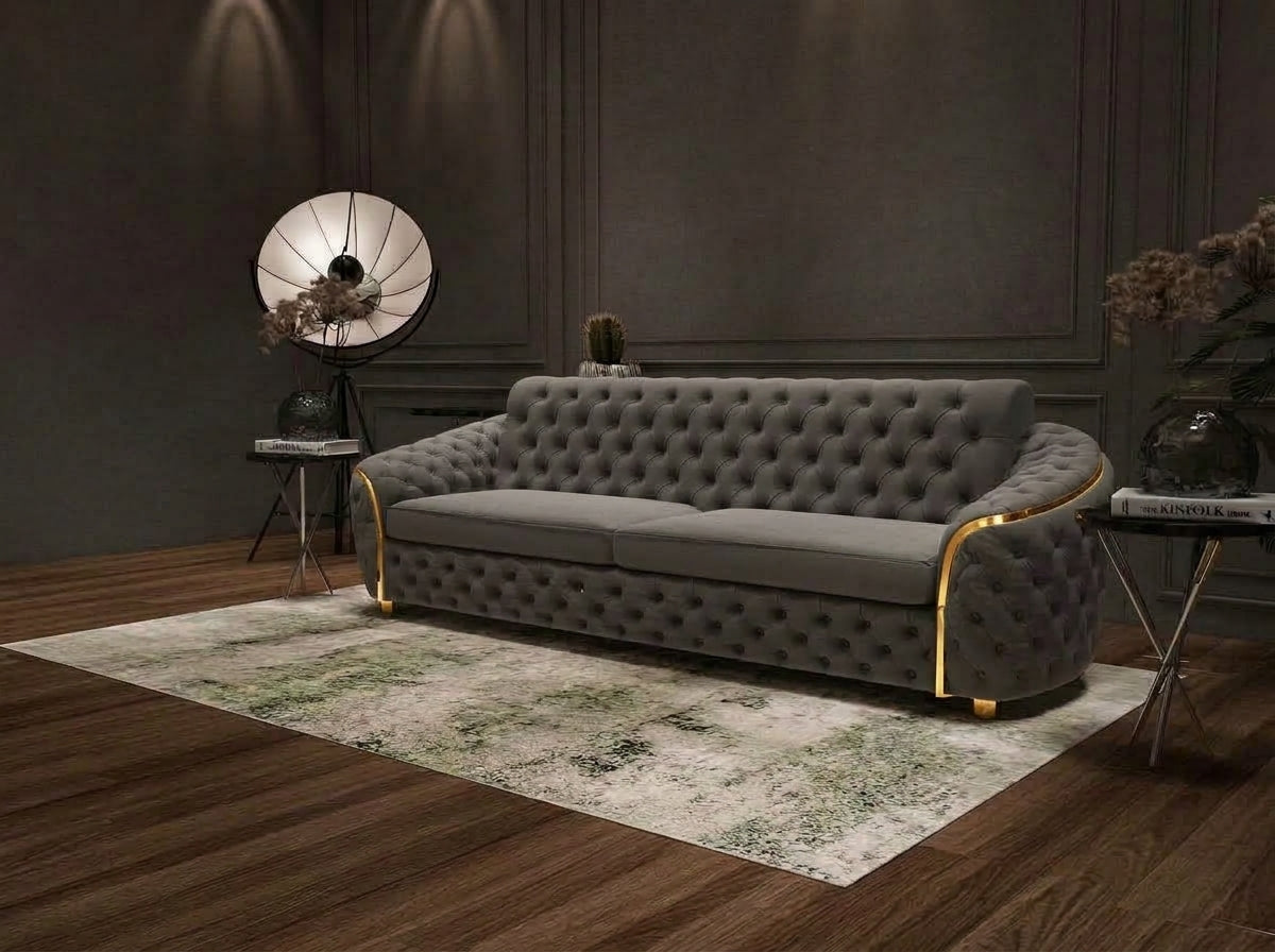 A modern tufted sofa in gray velvet with gold-toned metal accents, styled in a dark, elegant room on a distressed area rug.