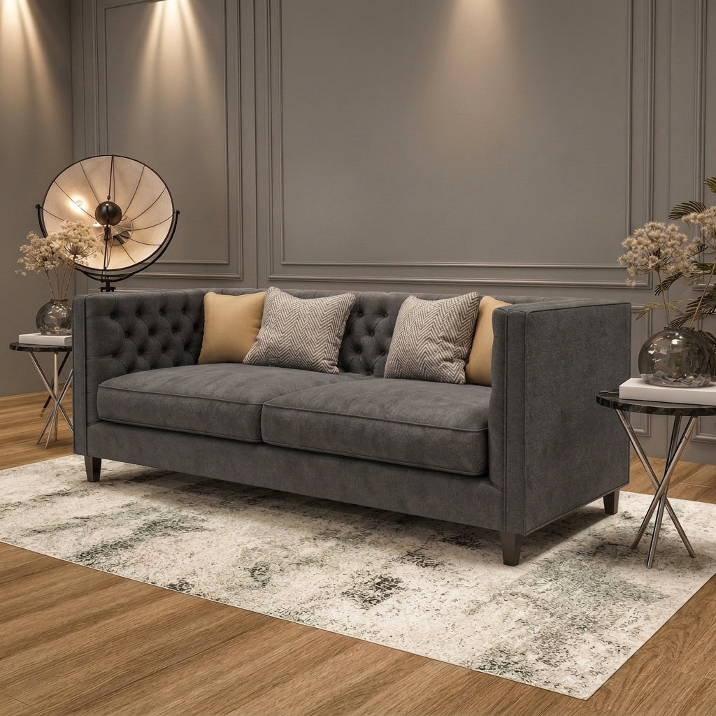 A modern gray fabric sofa with button tufting on the backrest, styled with three throw pillows in a contemporary living room setting with a distressed white rug, side tables, and a decorative lamp.